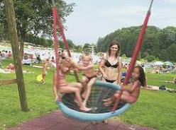 Vier Kinder sitzen und schwingen auf einer runden Schaukel in einem Park, während eine erwachsene Frau in der Nähe steht. Im Hintergrund sitzen Menschen im Gras und entspannen sich.