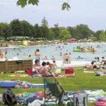 Menschen entspannen sich auf dem Rasen und schwimmen in einem überfüllten Freibad an einem sonnigen Tag, mit Handtüchern, Stühlen und Schwimmflügeln in Sichtweite.