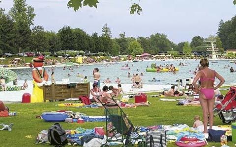 Menschen entspannen sich auf dem Rasen und schwimmen in einem überfüllten Freibad an einem sonnigen Tag, mit Handtüchern, Stühlen und Schwimmflügeln in Sichtweite.