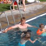 Ein Mann steht in einem Pool mit zwei Kindern, die orangefarbene Schwimmflügel tragen, während ein anderer Erwachsener in der Nähe sitzt. Im Hintergrund entspannen sich Menschen auf dem Rasen.