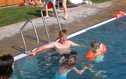 Ein Mann steht in einem Pool mit zwei Kindern, die orangefarbene Schwimmflügel tragen, während ein anderer Erwachsener in der Nähe sitzt. Im Hintergrund entspannen sich Menschen auf dem Rasen.
