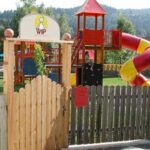 Ein eingezäunter Spielplatz mit einem "VIP"-Schild am Eingang, mit einer roten und gelben Spiralrutsche, Klettergerüsten und grünem Rasen.