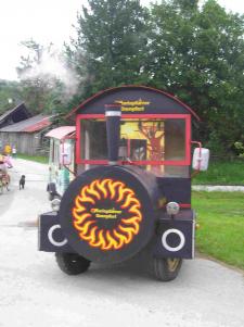 Ein kleines, buntes Fahrzeug in Form eines Zuges mit aufgemalten Flammen auf der Vorderseite ist auf einem gepflasterten Weg mit Bäumen und Gebäuden im Hintergrund geparkt.