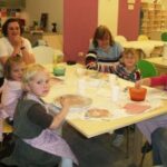 Mehrere kleine Kinder und ein paar Erwachsene sitzen in einem Klassenzimmer um einen Tisch herum und beschäftigen sich mit farbigen Materialien.