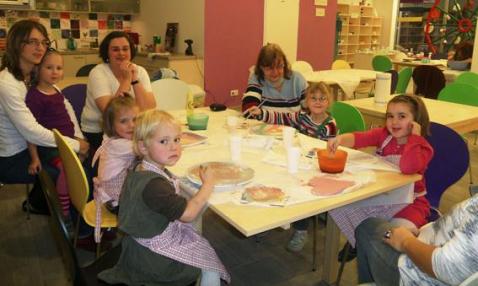 Mehrere kleine Kinder und ein paar Erwachsene sitzen in einem Klassenzimmer um einen Tisch herum und beschäftigen sich mit farbigen Materialien.