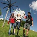 Zwei Erwachsene halten ein Kind an den Armen und schwingen es, während sie auf einem grasbewachsenen Hügel mit einer hölzernen Stangenkonstruktion und blauem Himmel im Hintergrund spazieren gehen.