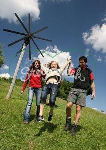 Zwei Erwachsene halten ein Kind an den Armen und schwingen es, während sie auf einem grasbewachsenen Hügel mit einer hölzernen Stangenkonstruktion und blauem Himmel im Hintergrund spazieren gehen.