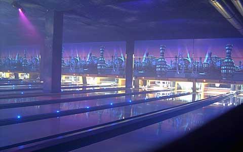 Eine Bowlingbahn mit leuchtenden blauen Lichtern, leeren Bahnen und futuristisch anmutender Wandmalerei im Hintergrund.