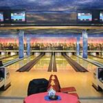 Vier Bowlingbahnen in einer hell erleuchteten Bowlingbahn mit elektronischen Anzeigetafeln über dem Kopf und Sitzbereichen im Vordergrund.