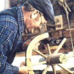 Ein Mann mit Mütze und kariertem Hemd arbeitet in einer Werkstatt am Zusammenbau eines hölzernen Wagenrads.