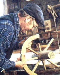 Ein Mann mit Mütze und kariertem Hemd arbeitet in einer Werkstatt am Zusammenbau eines hölzernen Wagenrads.