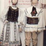 Zwei Schaufensterpuppen in traditioneller osteuropäischer Volkstracht mit gestickten Mustern und dekorativen Accessoires stehen vor einem floralen Wandhintergrund.