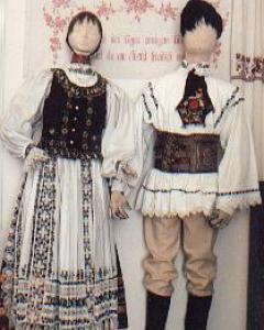 Zwei Schaufensterpuppen in traditioneller osteuropäischer Volkstracht mit gestickten Mustern und dekorativen Accessoires stehen vor einem floralen Wandhintergrund.