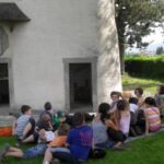 Eine Gruppe von Kindern sitzt im Gras vor einem weißen Gebäude und hört zwei Erwachsenen zu. Die Szene spielt sich an einem sonnigen Tag unter Bäumen ab.