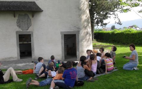 Eine Gruppe von Kindern sitzt im Gras vor einem weißen Gebäude und hört zwei Erwachsenen zu. Die Szene spielt sich an einem sonnigen Tag unter Bäumen ab.