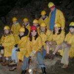 Eine Gruppe von Menschen in gelben Regenmänteln und Schutzhelmen sitzt und steht auf Felsen in einer schwach beleuchteten Höhle.