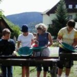 Fünf Personen stehen im Freien und wühlen mit grünen Pfannen nach Gold, während im Hintergrund Häuser und Berge zu sehen sind.