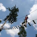 Eine Person mit einem Klettergurt überquert eine Hochseilgartenbrücke, die zwischen Bäumen vor einem blauen Himmel mit vereinzelten Wolken hängt.