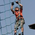 Ein Kind, das einen Helm und Handschuhe trägt, klettert im Freien bei klarem Himmel an einem blauen Seilnetz.