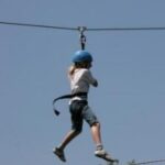 Eine Person, die einen blauen Helm und ein Gurtzeug trägt, fährt auf einer Seilrutsche gegen einen klaren Himmel.