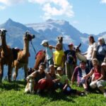 Eine Gruppe von Menschen, darunter auch Kinder, posiert mit mehreren Lamas auf einem grasbewachsenen Hügel mit Bergen und blauem Himmel im Hintergrund.