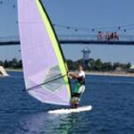 Eine Person beim Windsurfen auf einem Gewässer mit einer Brücke und mehreren Personen im Hintergrund an einem klaren, sonnigen Tag.
