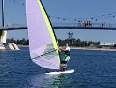 Eine Person beim Windsurfen auf einem Gewässer mit einer Brücke und mehreren Personen im Hintergrund an einem klaren, sonnigen Tag.