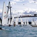 Mehrere kleine Segelboote fahren auf dem kabbeligen Wasser in der Nähe einer Fußgängerbrücke um die Wette, während die Menschen von der Brücke aus bei bewölktem Himmel zusehen.