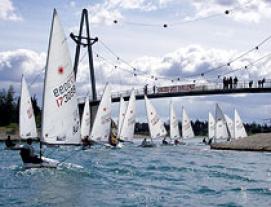 Mehrere kleine Segelboote fahren auf dem kabbeligen Wasser in der Nähe einer Fußgängerbrücke um die Wette, während die Menschen von der Brücke aus bei bewölktem Himmel zusehen.