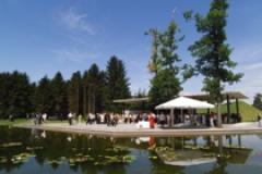 An einem sonnigen Tag versammeln sich Menschen unter Zelten an einem Teich mit Seerosenblättern, umgeben von Bäumen und Grasflächen.