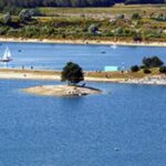Eine kleine Insel mit ein paar Bäumen ist von blauem Wasser umgeben, in der Nähe eines Ufers mit Booten, Menschen und einer Brücke im Hintergrund.