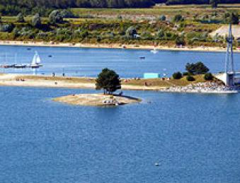 Eine kleine Insel mit ein paar Bäumen ist von blauem Wasser umgeben, in der Nähe eines Ufers mit Booten, Menschen und einer Brücke im Hintergrund.