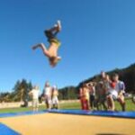 Ein Kind vollführt einen Rückwärtssalto auf einem Trampolin im Freien, während eine Gruppe von Kindern unter blauem Himmel zusieht.