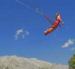 Eine Person in rotem Outfit macht einen Bungee-Sprung in der Luft vor einem blauen Himmel, im Hintergrund sind Berge und ein Baum zu sehen.