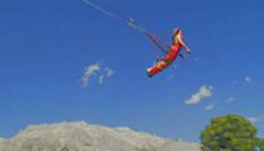 Eine Person in rotem Outfit macht einen Bungee-Sprung in der Luft vor einem blauen Himmel, im Hintergrund sind Berge und ein Baum zu sehen.