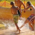 Drei Kinder in Badeanzügen planschen und spielen im flachen Wasser an einem sonnigen Tag im Freien, mit einem orangefarbenen Sicherheitszaun im Hintergrund.