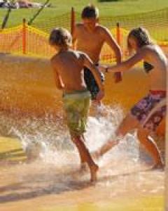 Drei Kinder in Badeanzügen planschen und spielen im flachen Wasser an einem sonnigen Tag im Freien, mit einem orangefarbenen Sicherheitszaun im Hintergrund.