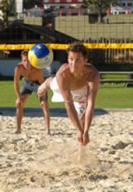 Zwei Männer spielen Beachvolleyball; ein Spieler taucht, um den Ball zu schlagen, während der andere hinter ihm auf einem Sandplatz steht.