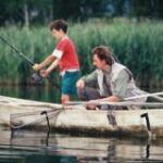 Ein Mann und ein Junge angeln von einem kleinen Holzboot aus auf einem ruhigen See, mit hohem Schilf im Hintergrund.