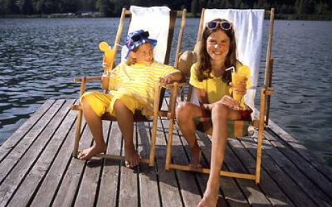 Zwei Kinder sitzen an einem sonnigen Tag auf Liegestühlen auf einem hölzernen Steg an einem See, lächeln und halten Eistüten in der Hand.