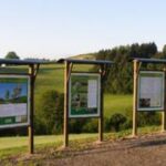 Drei Informationstafeln mit grüner und weißer Schrift stehen auf einem Kiesweg mit Blick auf grasbewachsene Felder und Bäume unter einem klaren Himmel.
