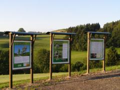 Drei Informationstafeln mit grüner und weißer Schrift stehen auf einem Kiesweg mit Blick auf grasbewachsene Felder und Bäume unter einem klaren Himmel.
