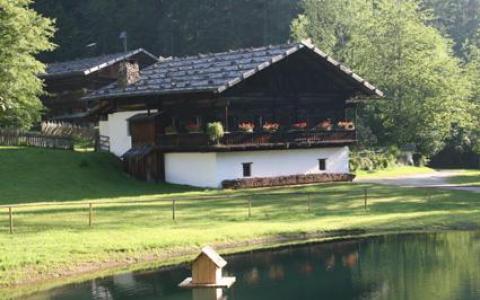 Ein rustikales Holzhaus mit Blumenkästen auf dem Balkon steht in der Nähe eines kleinen Teichs mit Zaun, umgeben von Gras und Bäumen.