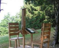 Zwei Holzstühle und ein kleiner Tisch sind im Freien aufgestellt; der Tisch hat einen ungewöhnlichen vertikalen Aufsatz, der ein Buch oder ein Tablet hält. Im Hintergrund sind Bäume und Grünflächen zu sehen.