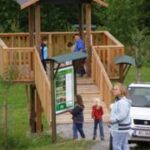 Mehrere Personen stehen in der Nähe einer hölzernen Aussichtsplattform und eines Informationsschildes in einem grasbewachsenen Außenbereich mit einem geparkten weißen Auto in der Nähe.