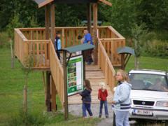 Mehrere Personen stehen in der Nähe einer hölzernen Aussichtsplattform und eines Informationsschildes in einem grasbewachsenen Außenbereich mit einem geparkten weißen Auto in der Nähe.