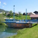 Ein hölzernes Spielgerät in Form eines Schiffes steht neben einem Teich, mit Gras, Häusern und Bergen im Hintergrund unter einem klaren Himmel.