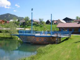Ein hölzernes Spielgerät in Form eines Schiffes steht neben einem Teich, mit Gras, Häusern und Bergen im Hintergrund unter einem klaren Himmel.