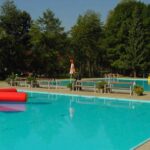 Freibad mit blauem Wasser, aufblasbaren Spielzeugen, Bänken am Beckenrand und einer gelben Rutsche; umgeben von Bäumen unter einem klaren Himmel.