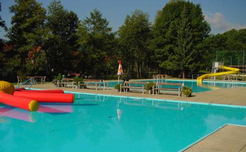 Freibad mit blauem Wasser, aufblasbaren Spielzeugen, Bänken am Beckenrand und einer gelben Rutsche; umgeben von Bäumen unter einem klaren Himmel.